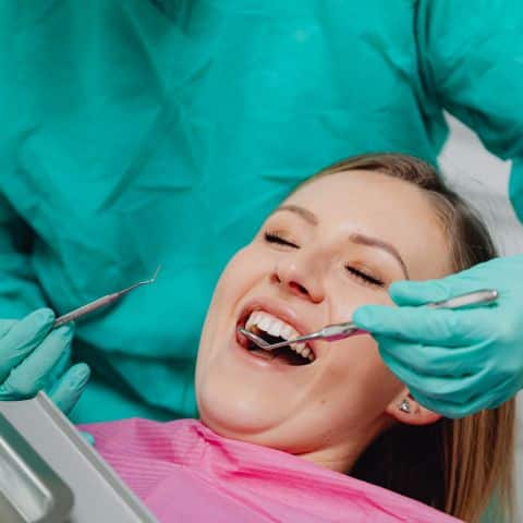 Paciente sorrindo durante avaliação odontológica, enquanto o dentista utiliza instrumentos para exame, ilustrando atendimento relacionado a Implante Zigomático.