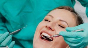 Paciente sorrindo durante avaliação odontológica, enquanto o dentista utiliza instrumentos para exame, ilustrando atendimento relacionado a Implante Zigomático.